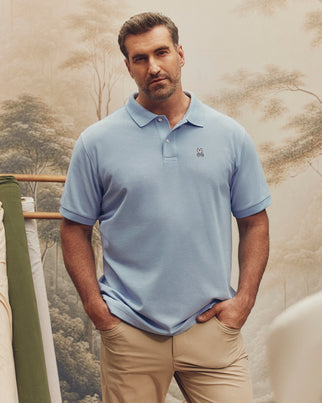 A man confidently stands with his hands in the pockets of his beige pants, wearing a light blue Big and Tall Classic Pique Polo - B9K001CRPC by Psycho Bunny, in front of a serene nature-themed backdrop with trees and soft lighting.