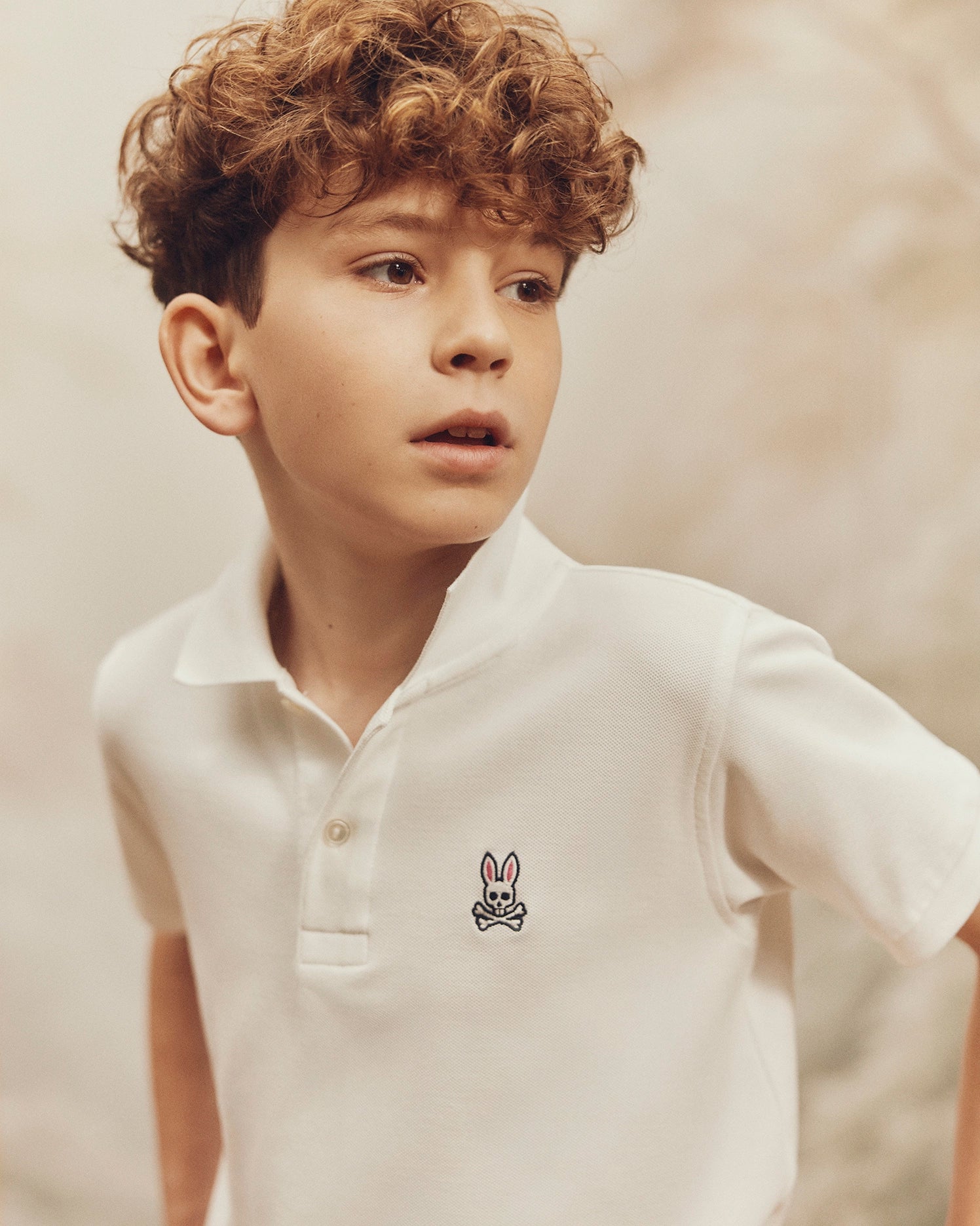 A boy with curly hair wears the Psycho Bunny KIDS CLASSIC PIQUE POLO SHIRT - B0K001CRPC, made of 100% Pima cotton with a small bunny emblem. He looks to the side in front of a softly blurred background, highlighting the shirt's mother-of-pearl buttons.