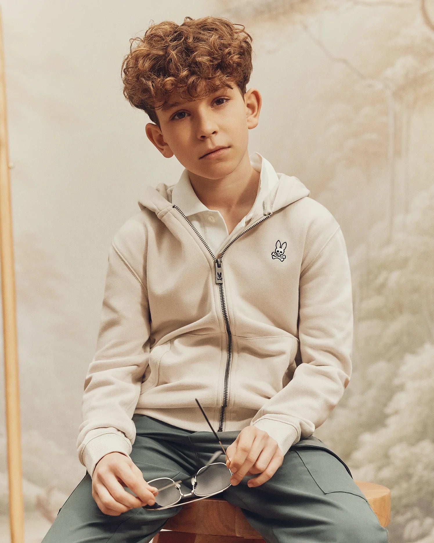 A young boy with curly brown hair sits on a wooden stool, wearing Psycho Bunny's KIDS LEVY ESSENTIAL ZIP FLEECE HOODIE and green pants. He's holding a pair of glasses in his hands. A soft, blurred nature background is behind him.