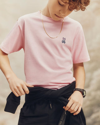 A young person with curly hair wears a pink Psycho Bunny Kids Classic Crew Neck Tee made from 100% Pima cotton, featuring a small rabbit logo. Paired with a gold chain necklace and dark jacket tied around their waist, it's perfect for back-to-school outfits, set against a softly blurred background.