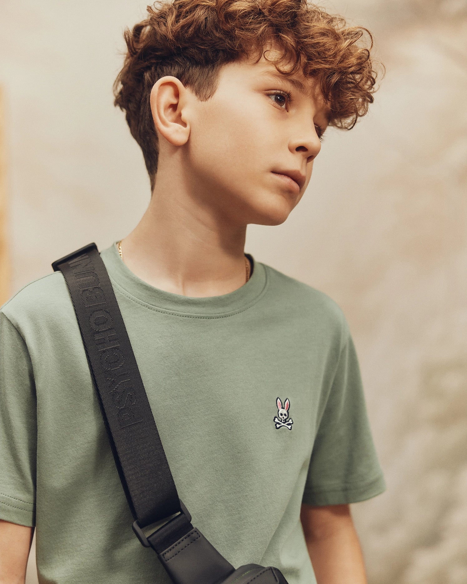 A young person with curly hair wears a Psycho Bunny KIDS CLASSIC CREW NECK TEE - B0U014B200, made from 100% Pima cotton, featuring a small bunny logo and carries a shoulder bag. They gaze thoughtfully as the blurred background creates a warm ambiance. Ideal for back-to-school outfits.