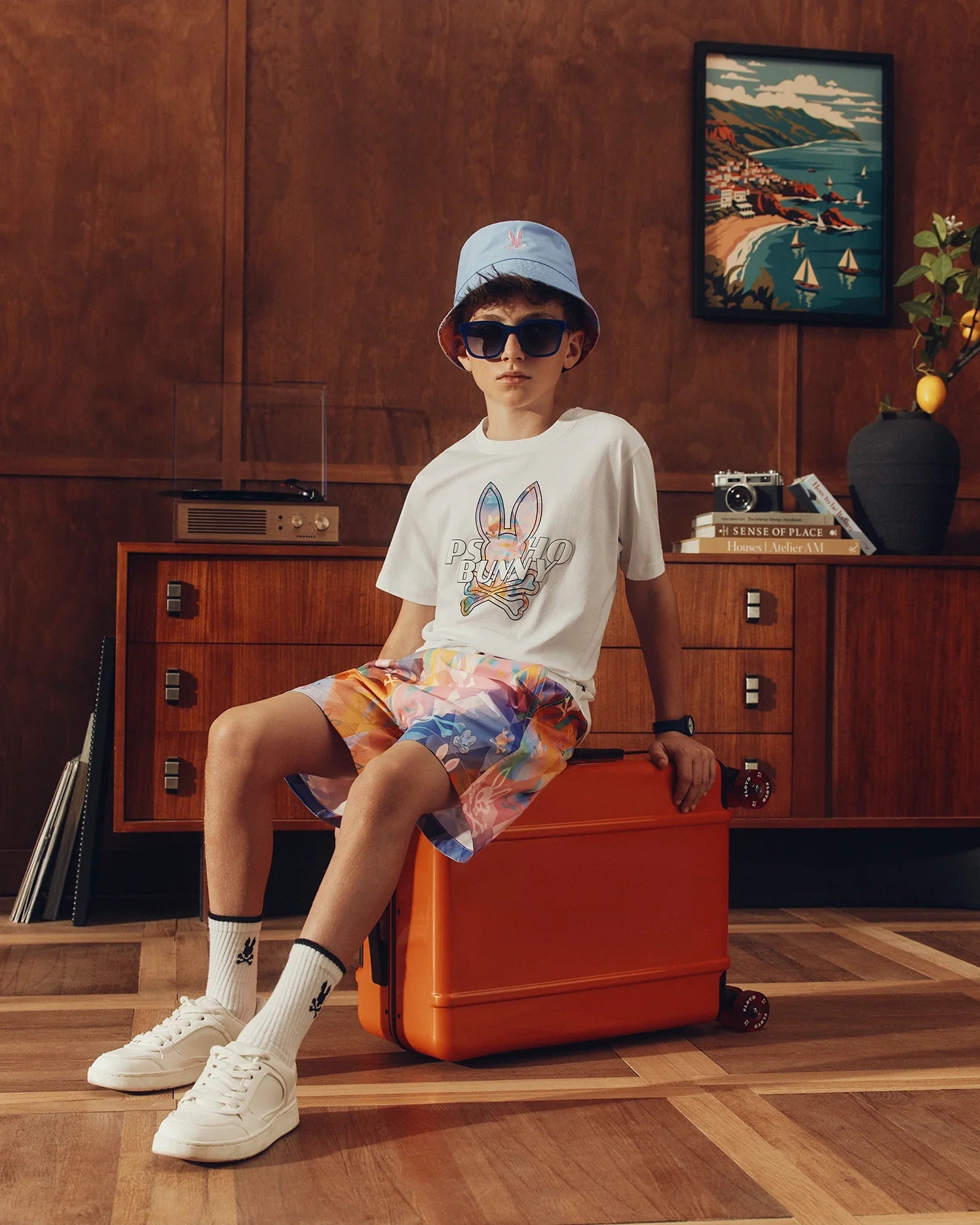 A young person sits on an orange suitcase in a stylish room, wearing a blue bucket hat and sunglasses. They're in the 