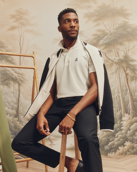 A man sits on a wooden stool wearing a Psycho Bunny MENS CLASSIC PIQUE POLO SHIRT - B6K001ARPC, black pants, and a jacket over his shoulders. The background has a tree mural and part of a wooden clothing rack is visible.