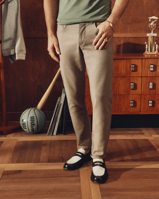 A person in Psycho Bunny's MENS MAVERICK 10K PANT - B6P788D200 and black-and-white loafers stands in a stylish room. A basketball and baseball bat lean against a wooden cabinet, topped by a trophy.