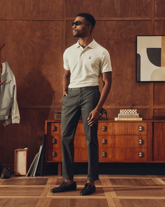 A stylish man wearing sunglasses stands confidently in a wooden-walled room, dressed in a white polo and Psycho Bunny's MENS MAVERICK 10K PANT - B6P788D200, celebrated for its five-pocket functionality and four-way stretch. A mid-century dresser with books and art sits behind him.