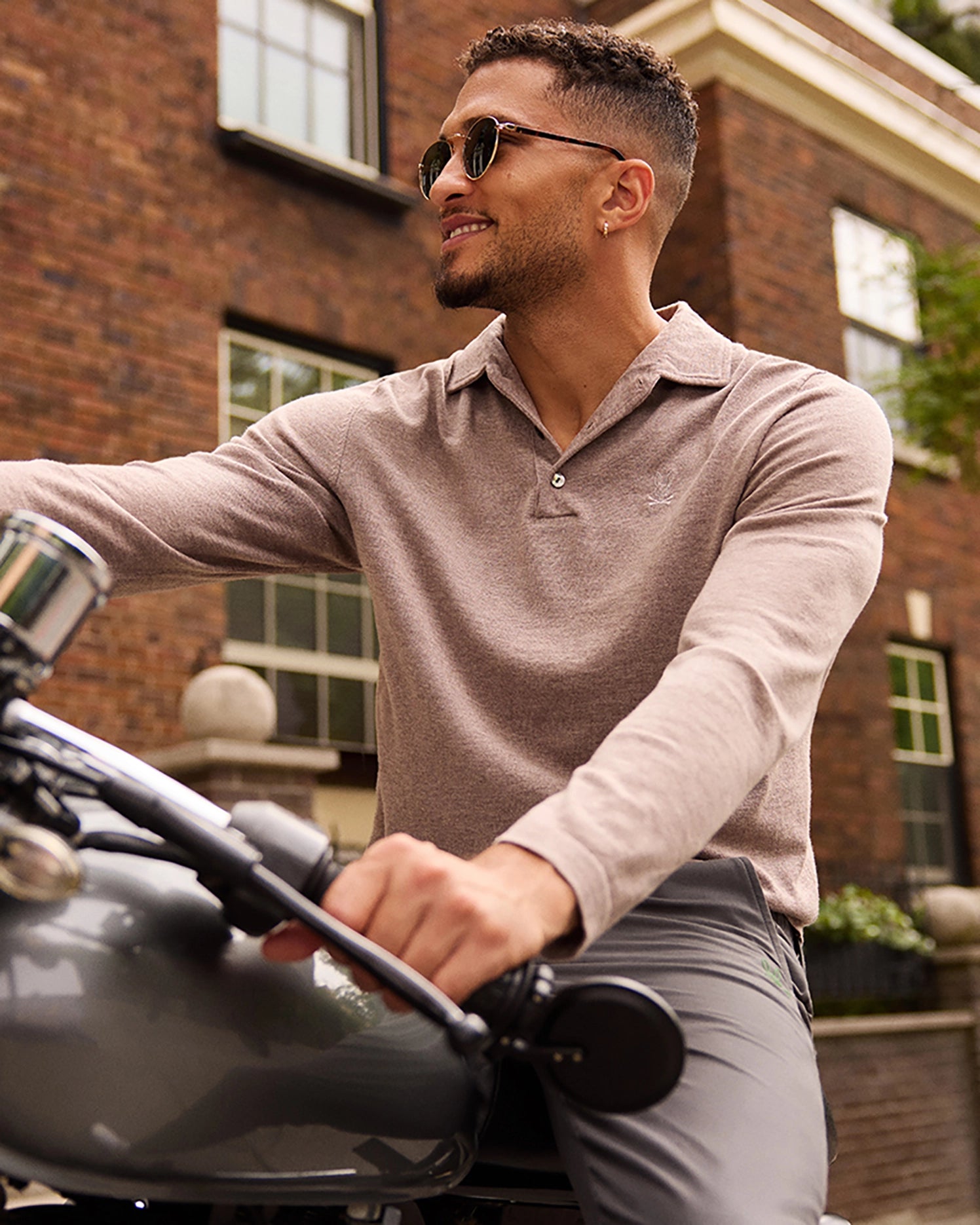 A man with short curly hair and sunglasses is smiling while sitting on a motorcycle. He is wearing the Psycho Bunny MENS BRADSHAW POLO SWEATER - B6E835D200, a beige long-sleeve shirt made from a soft and comfortable merino wool polo blend, along with gray pants. The background features a brick building with windows and some greenery.