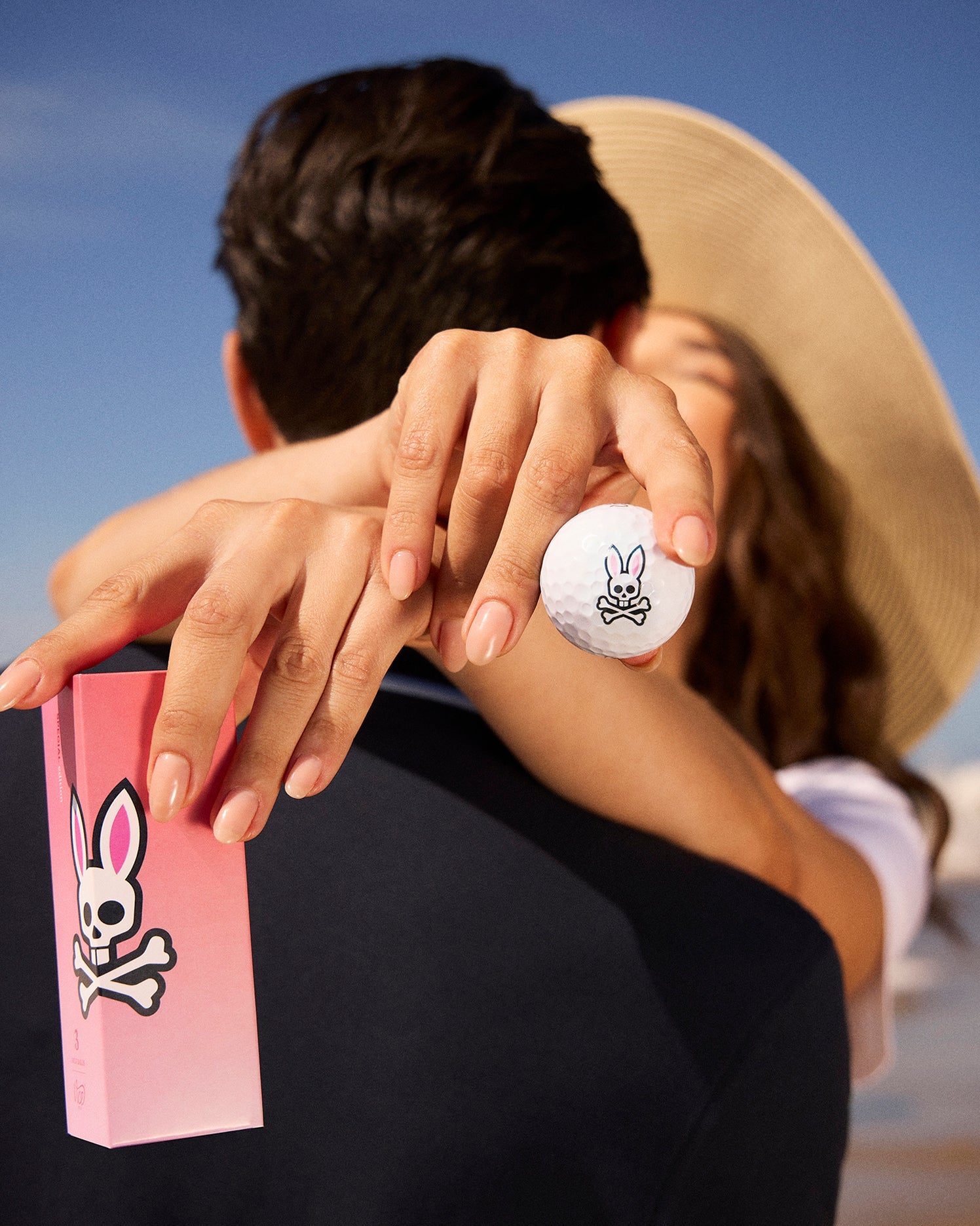 A woman in a sun hat embraces a man from behind, holding a Psycho Bunny 3 PACK GOLF BALLS - B6A565C200 with bunny ears in one hand and a matching pink box in the other. The pair is outdoors on a sunny day, and the man's face is mostly obscured by the woman's arms, hinting at their whimsical Vice collaboration.