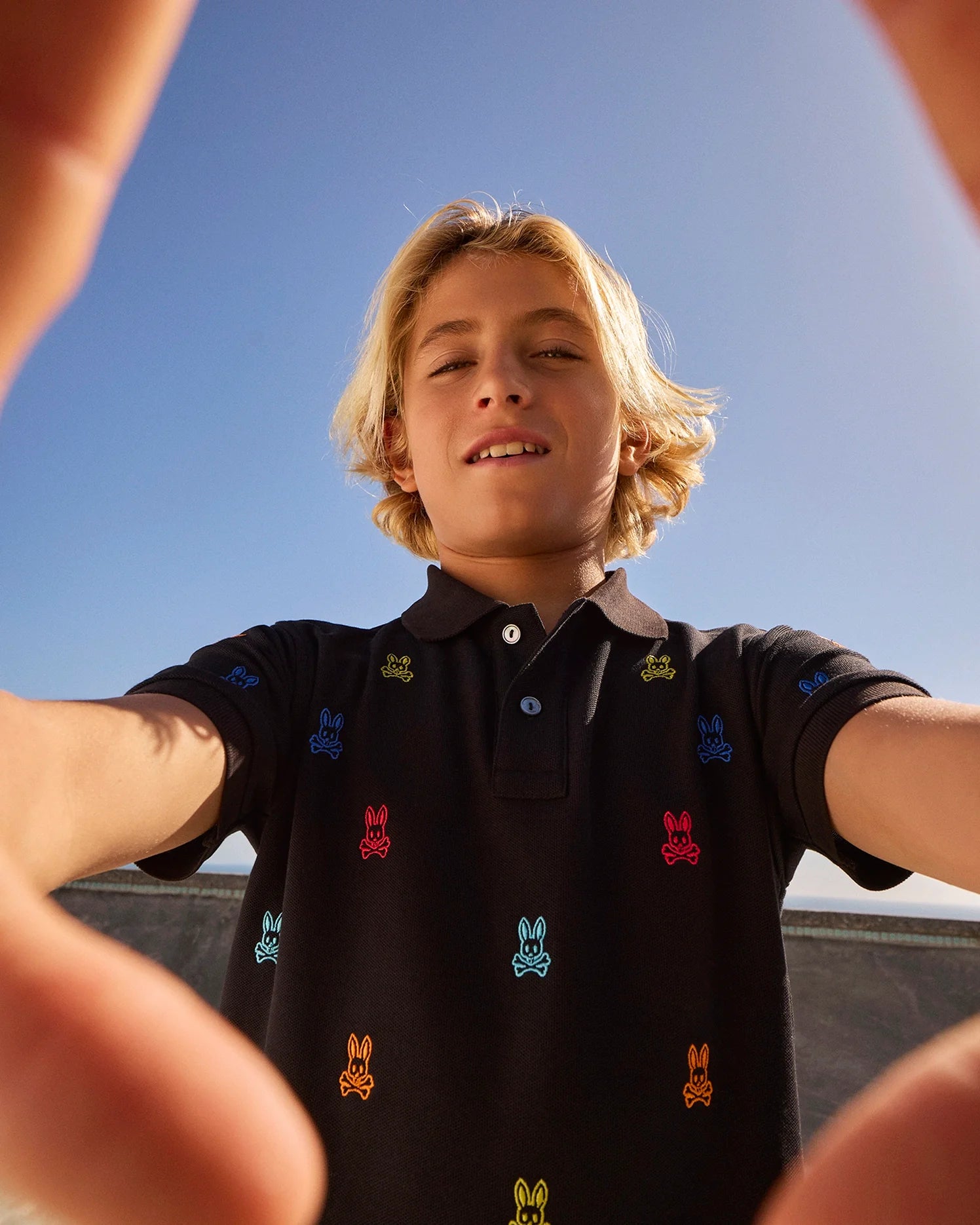 Seen from a low angle and framed between their hands, a young person with blond hair looks directly into the camera. They are wearing the Psycho Bunny KIDS BONHAM ALL OVER BUNNY PIQUE POLO (B0K501C200), which is black and adorned with colorful embroidered bunny motifs, set against a clear blue sky background.