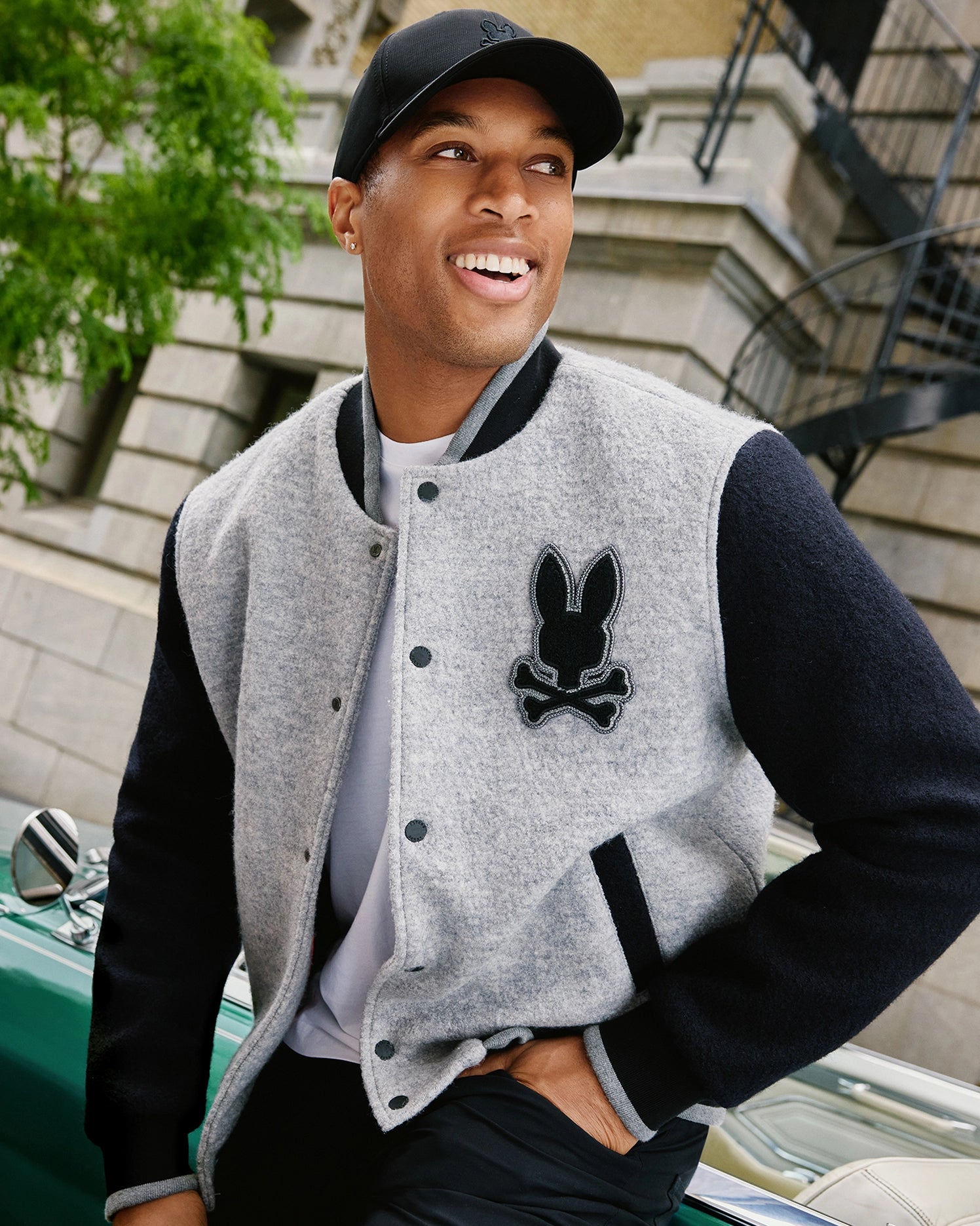 A man wearing a black baseball cap and the MENS TOM WOOL BOMBER JACKET - B6J744D200 by Psycho Bunny, featuring a chenille bunny patch, is smiling and leaning back against a green classic car. He is outdoors in an urban setting with a brick building and fire escape in the background.