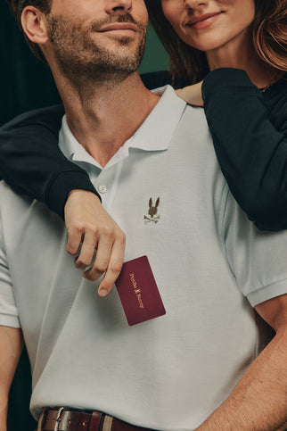 A man in a white Polo shirt eager for his holiday presents. Next to him, a woman in a black top wraps one arm around his shoulder while holding onto a Psycho Bunny Digital Gift Card—a perfect token of choice for their journey.
