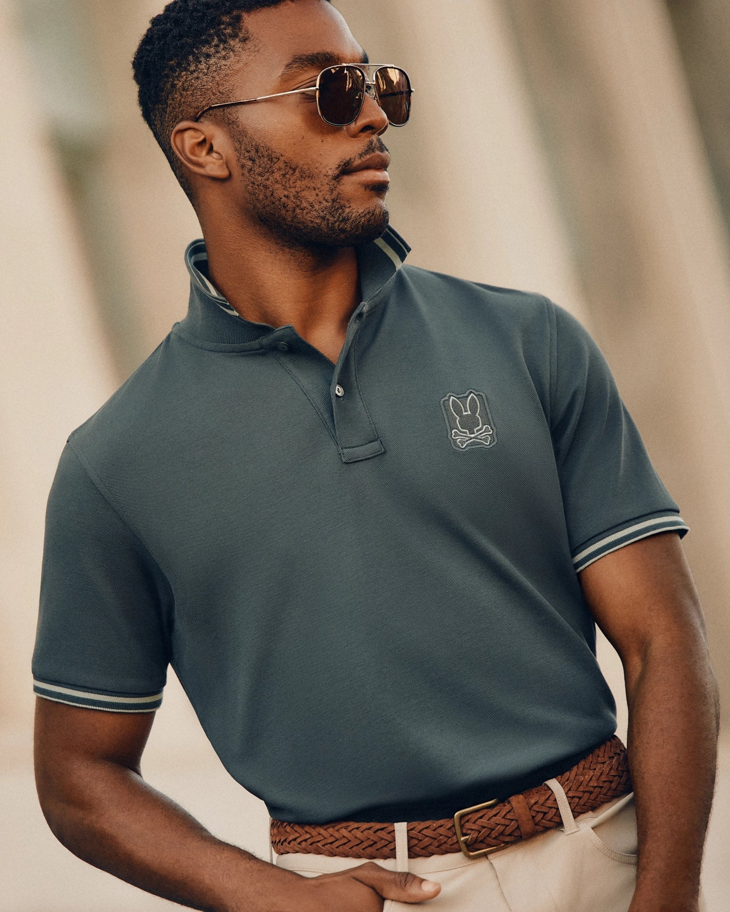 A confident man with a neatly trimmed beard is wearing sunglasses and the Psycho Bunny's MENS PIERCE PIQUE POLO (B6K201E200) in a dark green shade, featuring an embroidered Bunny logo. He has paired the polo with a woven brown belt and light pants against a softly blurred background.