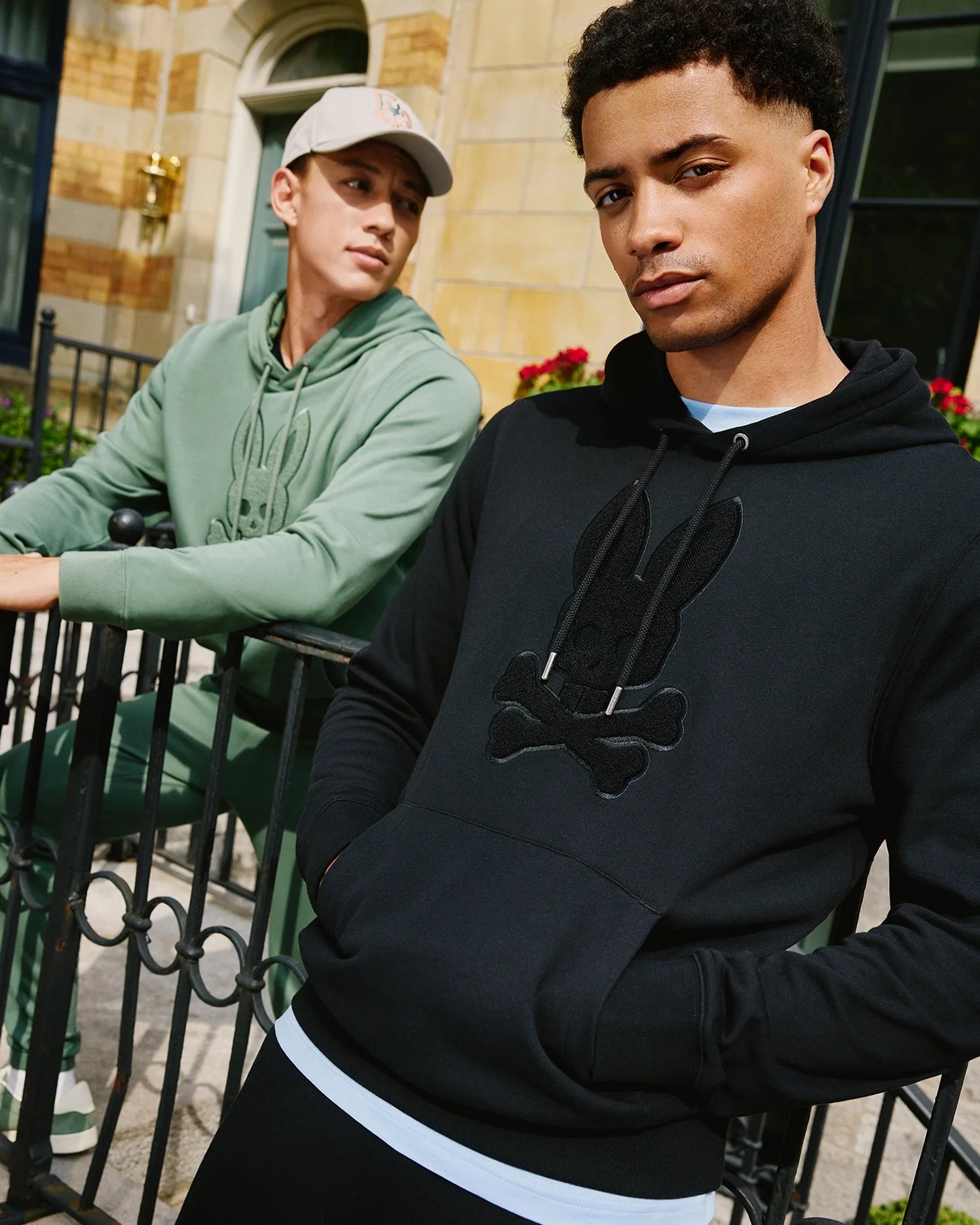 Two young men are standing outside in front of a building. The man in the foreground is wearing the Psycho Bunny MENS PACIFIC CHENILLE HOODIE - B6H930D200, featuring a black French terry fabric with a rabbit and crossbones chenille embroidery patch, while the man in the background is wearing a green 100% cotton hoodie and a beige cap. Both are leaning on a wrought iron fence.