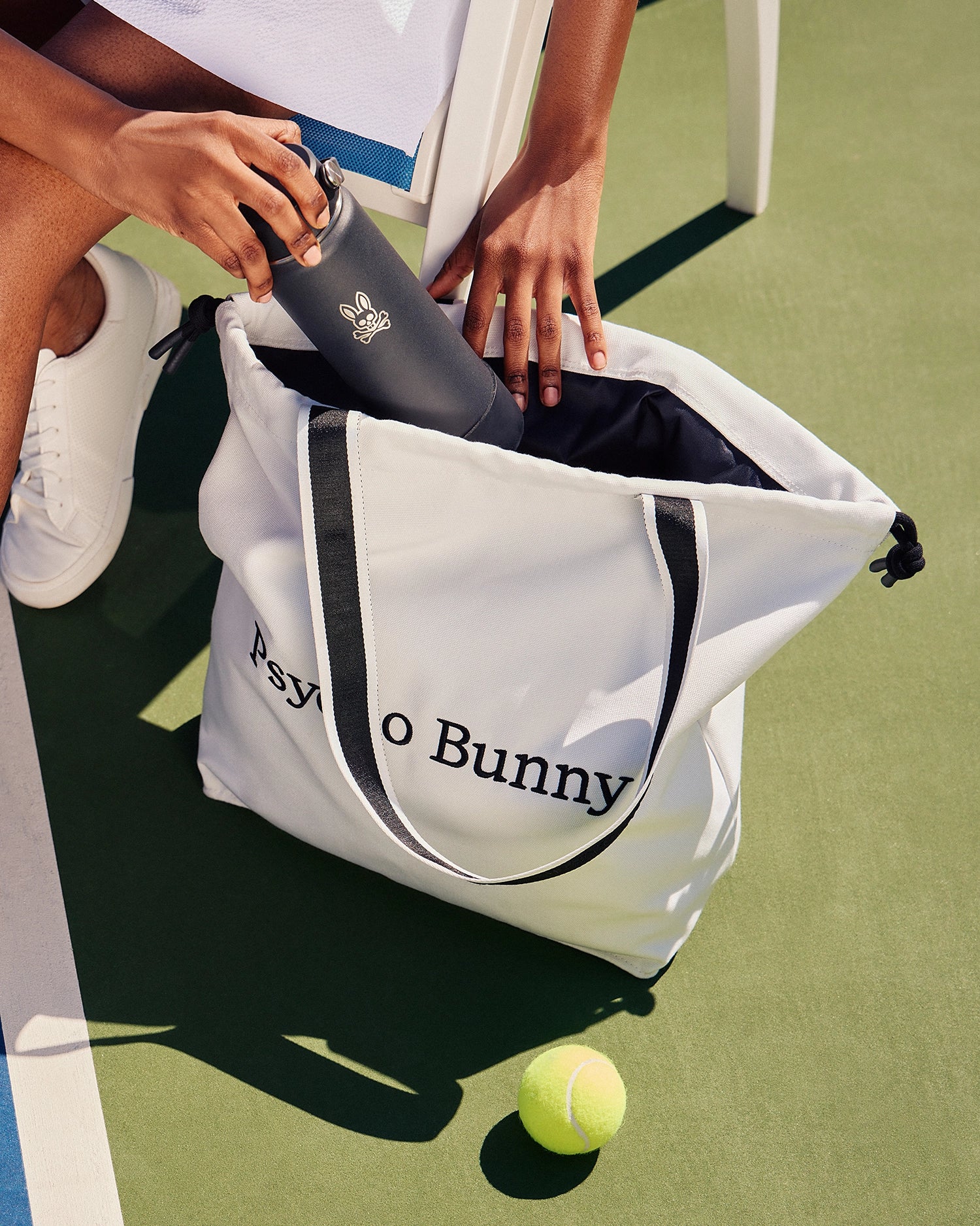 A person places a black water bottle into the luxurious TENNIS TOTE - B6A596C200 canvas bag on the court. The tote, featuring jacquard-woven straps and the 