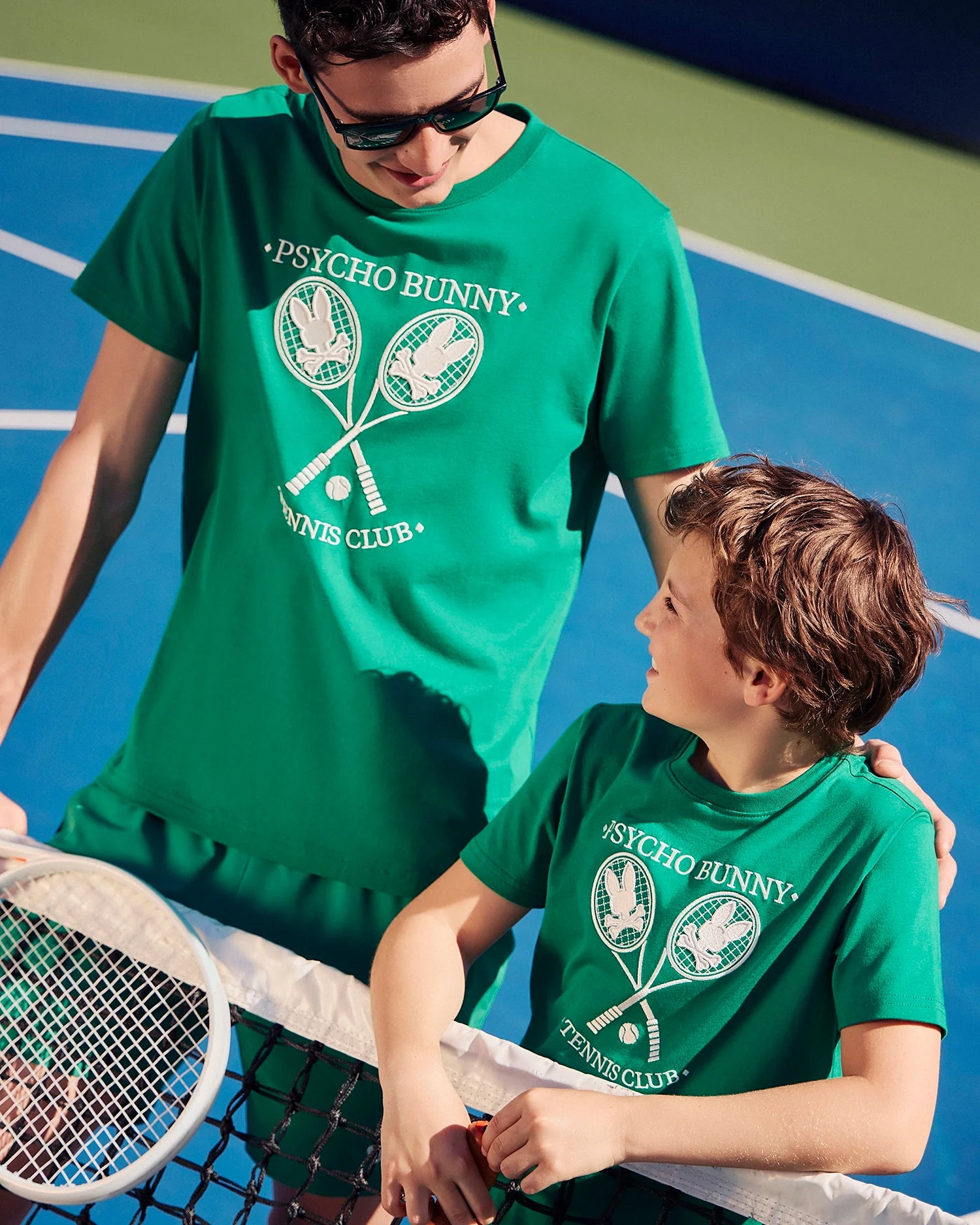 A man and a child stand on a tennis court, both wearing matching green Psycho Bunny Men's Courtside Graphic Tee - B6U684C200 Pima cotton jersey T-shirts. They are smiling at each other, with the man holding a tennis racket. The sunny court features a blue playing surface and green surroundings, enhancing the tennis-inspired scene.