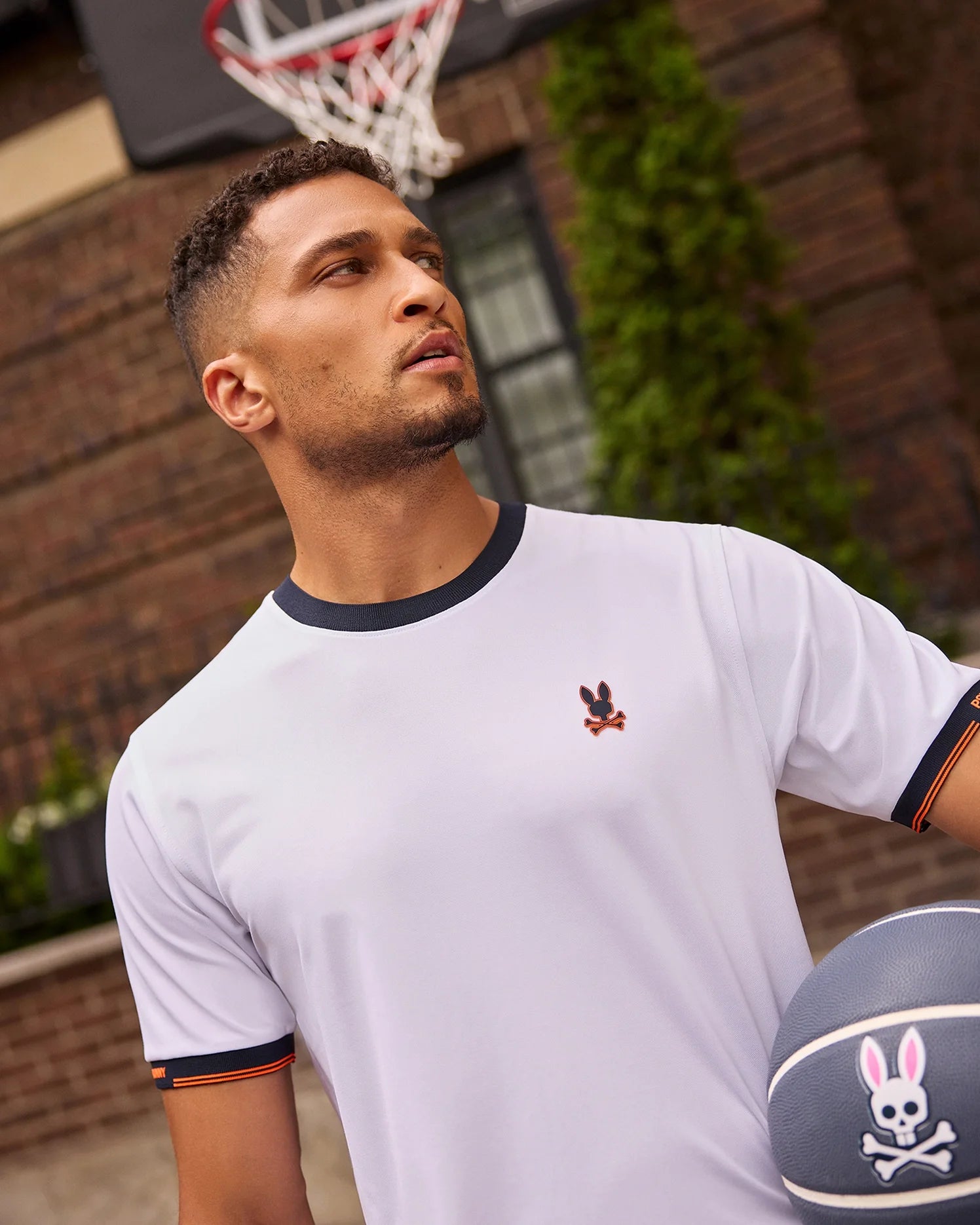 A man wearing the Psycho Bunny MENS EDMONDS SPORT TEE - B6U857D200, featuring a white base with black edges and a black and pink bunny logo, is standing on a basketball court. He holds a basketball that displays the same bunny logo. The background includes a brick building, a window, and a basketball hoop. This performance-oriented shirt is designed with antimicrobial properties to enhance breathability.