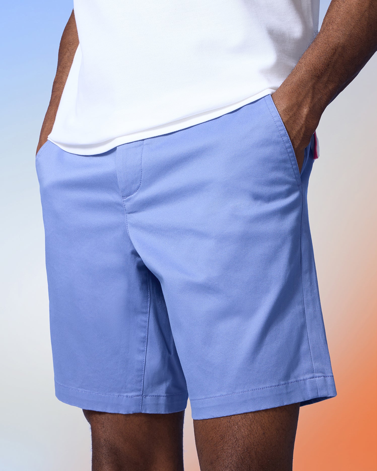 A person is wearing Psycho Bunny MENS YORK CHINO SHORT - B6R357B200 paired with a white shirt. The shorts have pockets, and the person's hands are casually placed in them. The background features a gradient effect with shades of blue and peach.