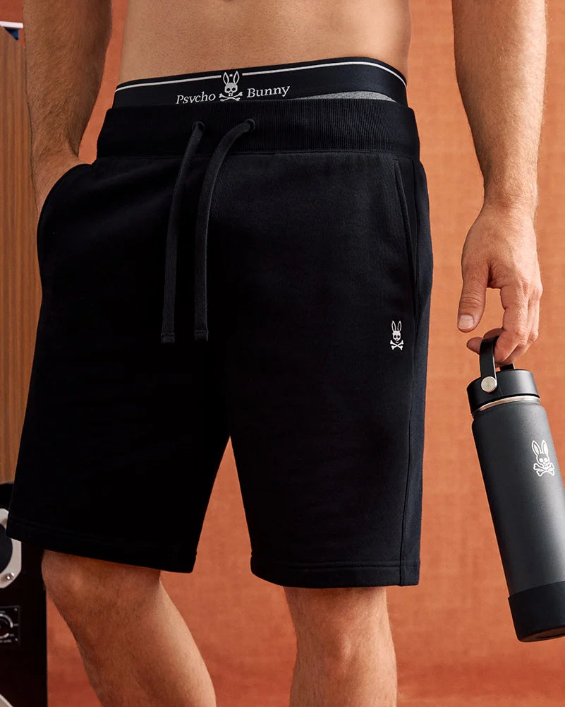 A person wearing Psycho Bunny's regular fit MENS LEVY FLEECE SWEATSHORT (B6R862D200) in black with an elastic drawstring waistband and a soft cotton fleece fabric, featuring a small rabbit logo on the left leg, holds a black water bottle in their right hand. Only the lower torso is visible.