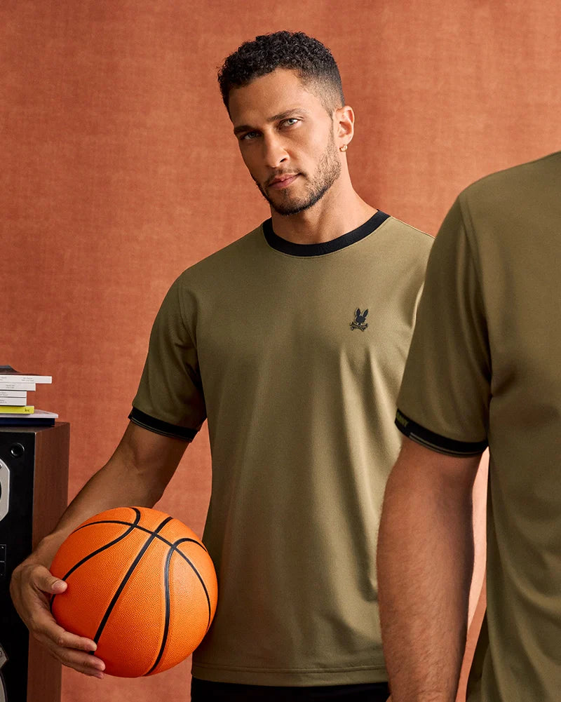 A man with short, curly hair and a beard is holding an orange basketball in his right hand. He is wearing the MENS EDMONDS SPORT TEE - B6U857D200 by Psycho Bunny, an olive-green T-shirt with black trim on the collar and sleeves, known for its breathability and quick-drying features. The background features a stack of books and a speaker against a brownish backdrop.