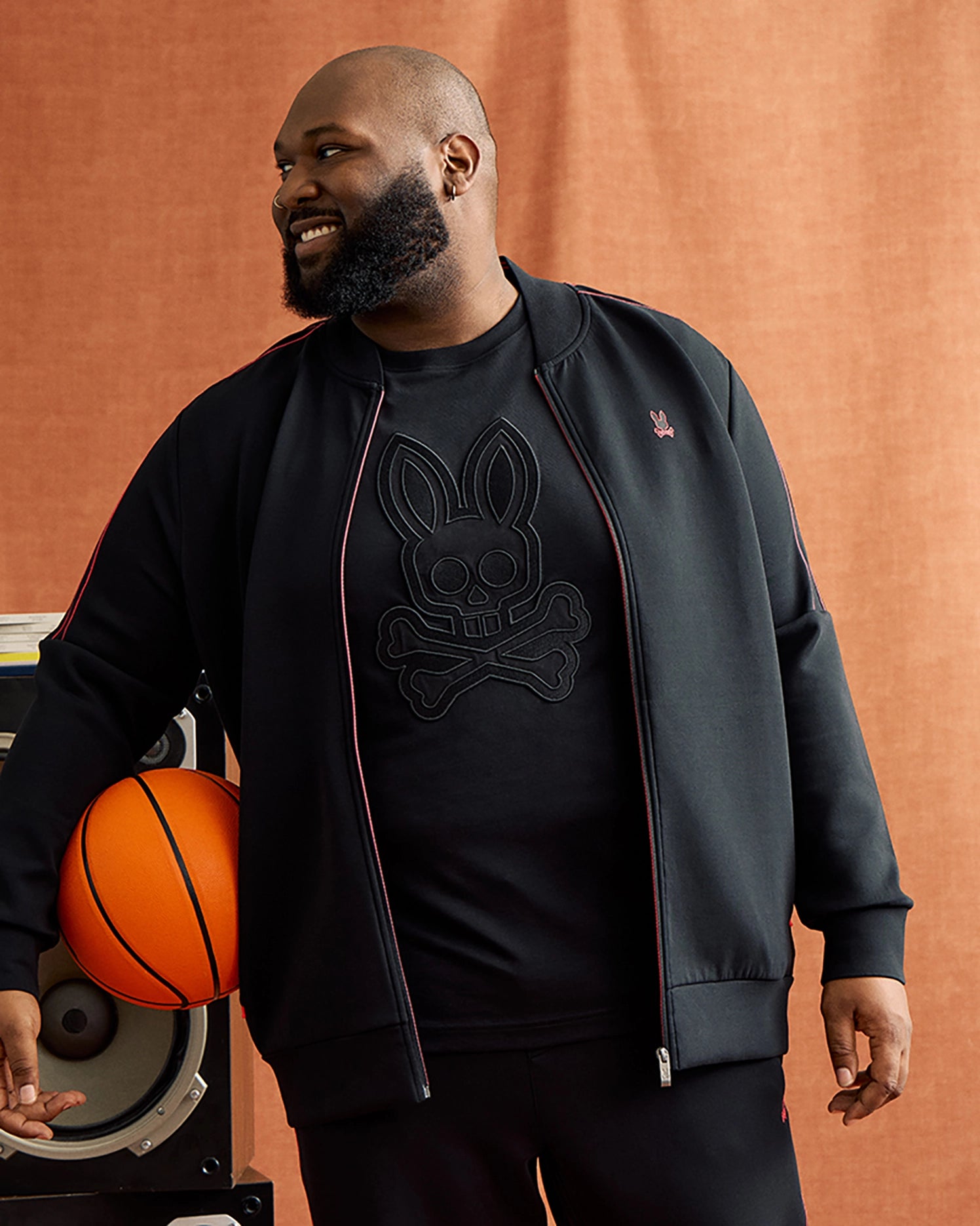 A bearded man, smiling and glancing to the side, dons the Psycho Bunny BIG AND TALL TRAVIS TRACK JACKET (B9J142D200), distinguished by its contrast chainstitch stripes. Underneath, he sports a black shirt adorned with a skull and crossbones motif. One of his hands rests on a shelf holding a basketball against a light brown background.