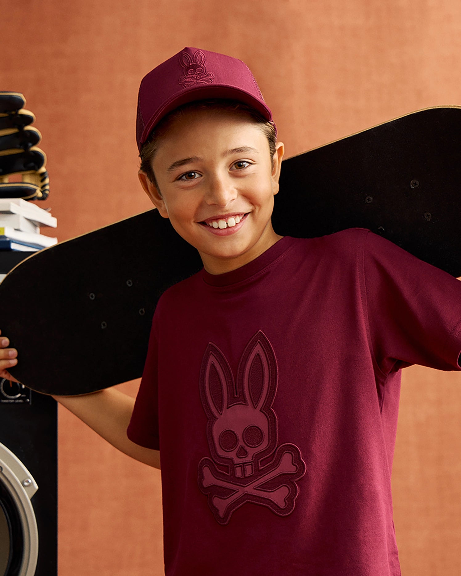 A smiling child wearing a maroon cap and a comfortable KIDS MALTON GRAPHIC TEE - B0U795D200 by Psycho Bunny, featuring a bunny with crossbones design, carries a skateboard over their shoulders. The background is a warm, textured orange.