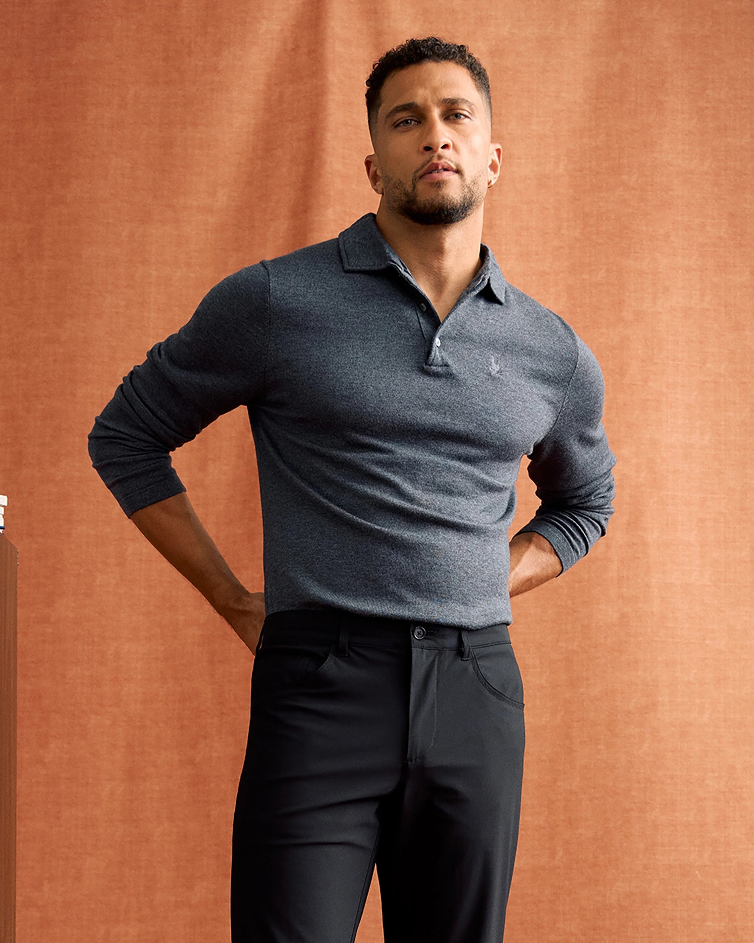 A man stands confidently against a rust-colored backdrop, donning a charcoal gray Merino wool MENS BRADSHAW POLO SWEATER - B6E835D200 by Psycho Bunny and black trousers. His hands rest on his hips, and he wears a slight smile. The luxury men's polo exudes sophistication and charm.