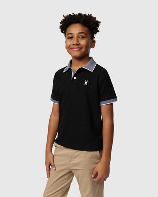 A young person with curly hair is smiling while wearing a Kids Southport Pique Polo Shirt (B0K263B200) by Psycho Bunny in black with grey trim, paired with beige pants. The Pima cotton shirt features a small embroidered logo on the left side against a plain, light-colored background.