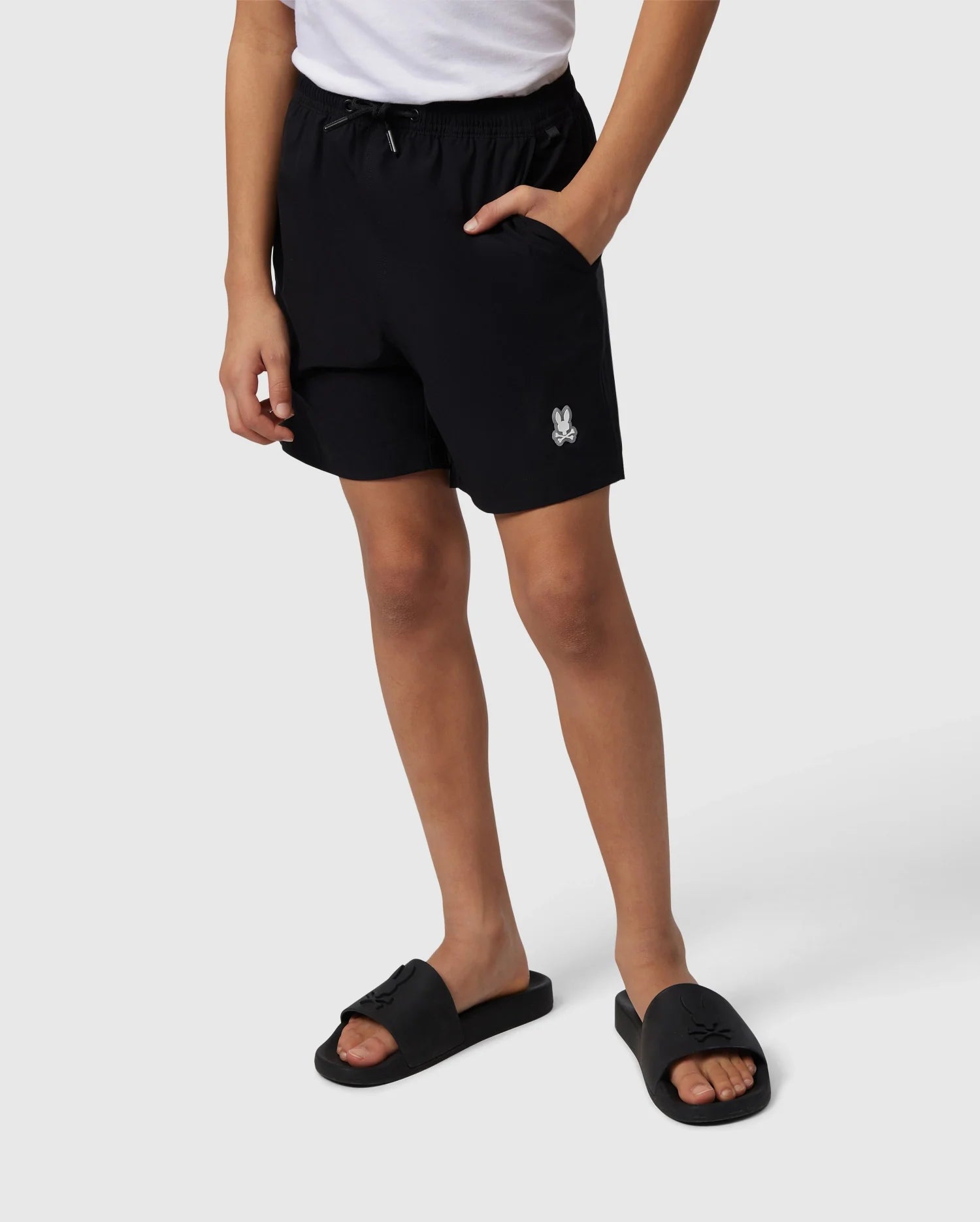 A person is shown from the waist down, wearing Psycho Bunny's KIDS MALTA HYDROCHROMIC SWIM TRUNK - B0W321B2SW in black, featuring a white rabbit logo on the left leg, and black slide sandals. They have their right hand in the pocket of the shorts, standing against a plain white background. The bunny pattern on the swim trunks hints at a playful touch.