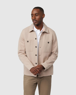 A person stands against a plain background, wearing an elevated style beige men's Bedford shirt jacket from Psycho Bunny (product code: B6J290B200) over a white collared shirt and khaki pants. This jacket, celebrated for its exceptional on-body feel, features two buttoned chest pockets. The person looks slightly to the side with hands gently clasped in front.
