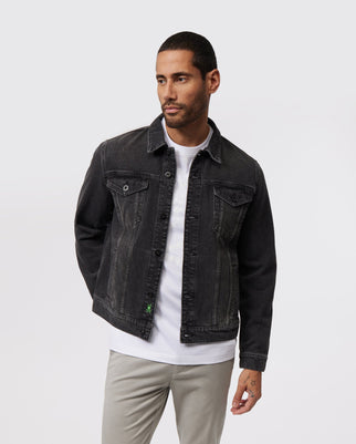 A man with short hair and a beard is wearing the Psycho Bunny MENS PATHE DENIM JACKET - B6J741X1CO with adjustable cuffs over a white t-shirt and light gray pants. He stands against a plain white background, hands in pockets, embodying the essence of men's fashion as he gazes to his left.