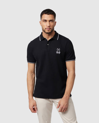 A man stands against a plain gray background, wearing a black Psycho Bunny MENS LENOX PIQUE POLO SHIRT - B6K138B200 with white trim on the collar and sleeves. The shirt features mother-of-pearl buttons and has a small embroidered bunny icon on the left side of the chest. He completes his outfit with light-colored pants.