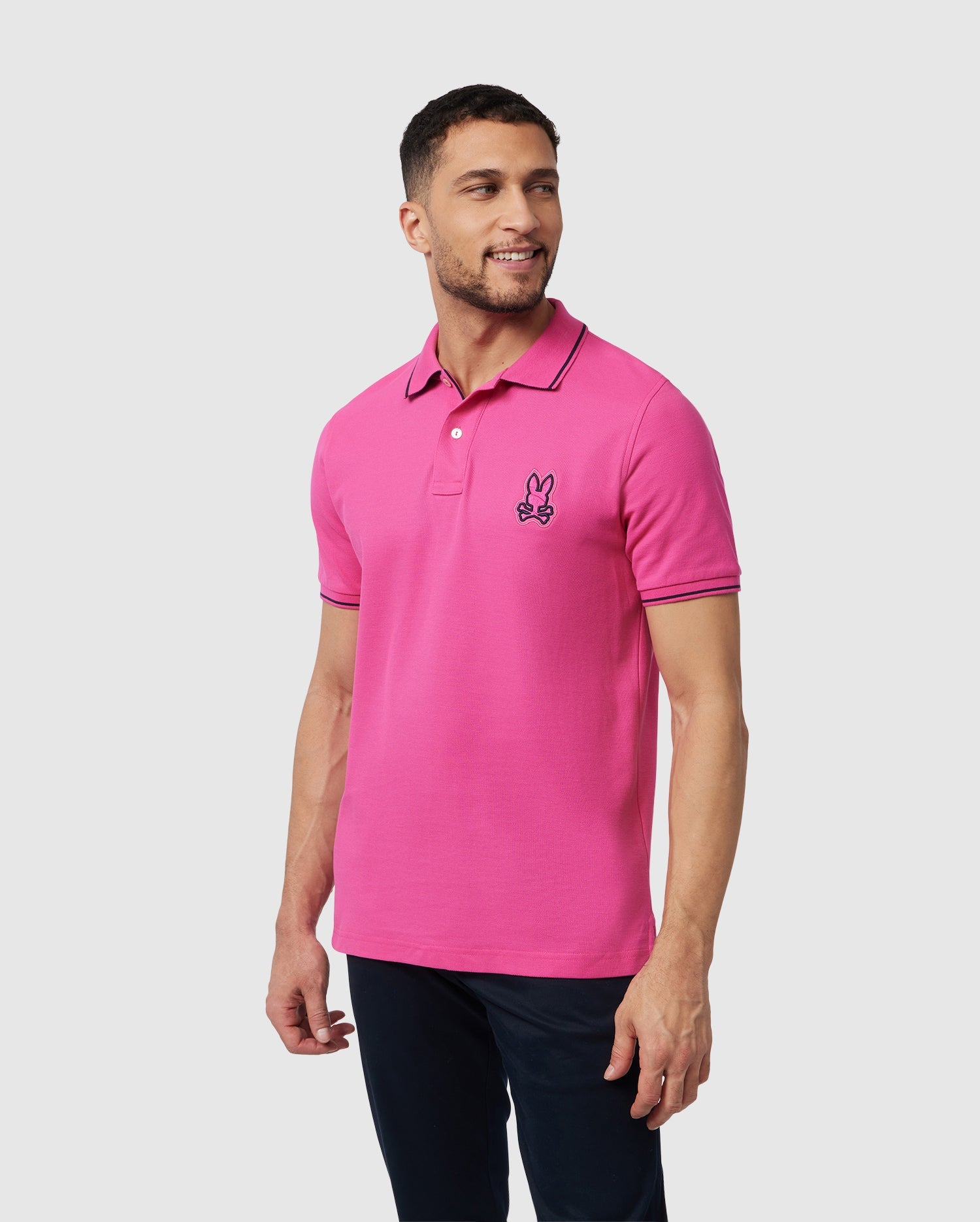 A man with short hair and a beard wears a pink Psycho Bunny MENS LENOX PIQUE POLO SHIRT - B6K138B200 made of soft Pima cotton, featuring an embroidered Bunny logo on the chest. He is looking to his right and is smiling slightly. The background is plain white.