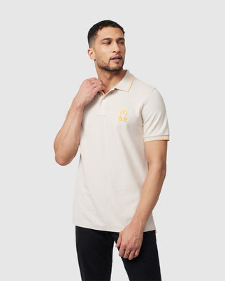 A person with short hair is wearing a cream-colored Psycho Bunny MENS LENOX PIQUE POLO SHIRT - B6K138B200 with a yellow embroidered Bunny logo on the left chest. They are standing against a plain light gray background, with one hand resting on their neck and the other by their side.