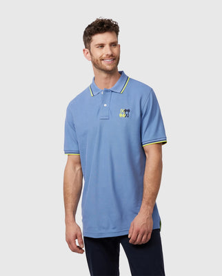 A man with short brown hair and a beard is wearing a light blue MENS WINTON PIQUE POLO SHIRT - B6K841A2PC by Psycho Bunny made of Peruvian Pima cotton with an embroidered Bunny design on the left chest. He is also wearing dark pants and is standing against a plain, light gray background. He is smiling and looking slightly off to the side.