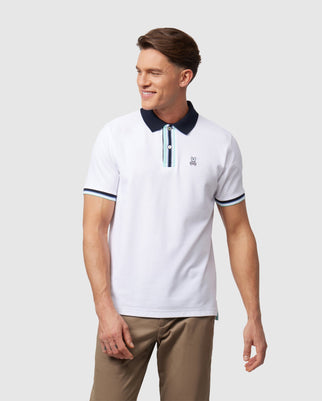 A man is wearing a Psycho Bunny MENS BLOOMINGTON PIQUE POLO SHIRT - B6K900A2PC made from Peruvian Pima cotton, featuring a white body with a navy blue collar and sleeve trim. The shirt has a small embroidered logo on the chest and a striped placket under the buttons. He stands against a plain background, smiling and looking to the side.