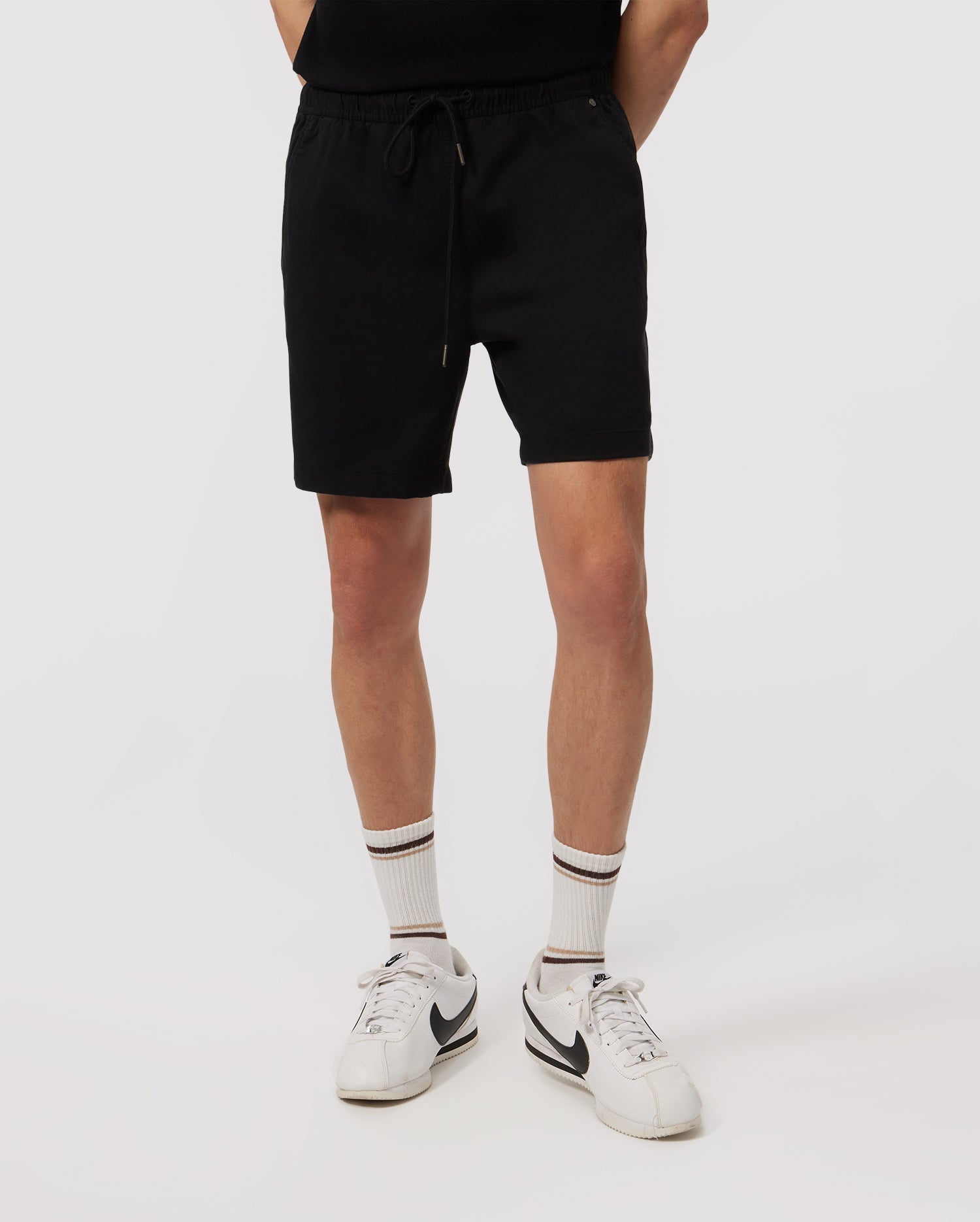 A person stands with their hands behind their back, wearing Psycho Bunny MENS WILLIS STRETCH TENCEL SHORT - B6R239Y1WB, white socks with black stripes, and white sneakers. The background is plain white. The photo is cropped at the waist and mid-shin, not showing the person's face or upper body.