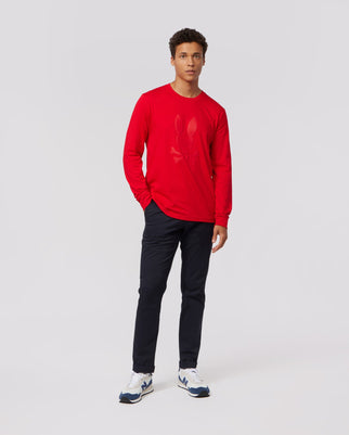 A person stands against a plain background wearing a textured red Psycho Bunny MENS SACRAMENTO LONG SLEEVE TEE - B6T695A2PC, black pants, and white sneakers. They have a neutral expression with their hands at their sides.