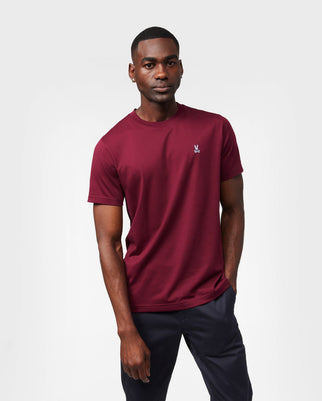 A person wearing a Psycho Bunny B6U014Z1PC ultra-soft Pima cotton men's classic crewneck tee in maroon with a small logo on the chest and navy pants is standing against a white background.