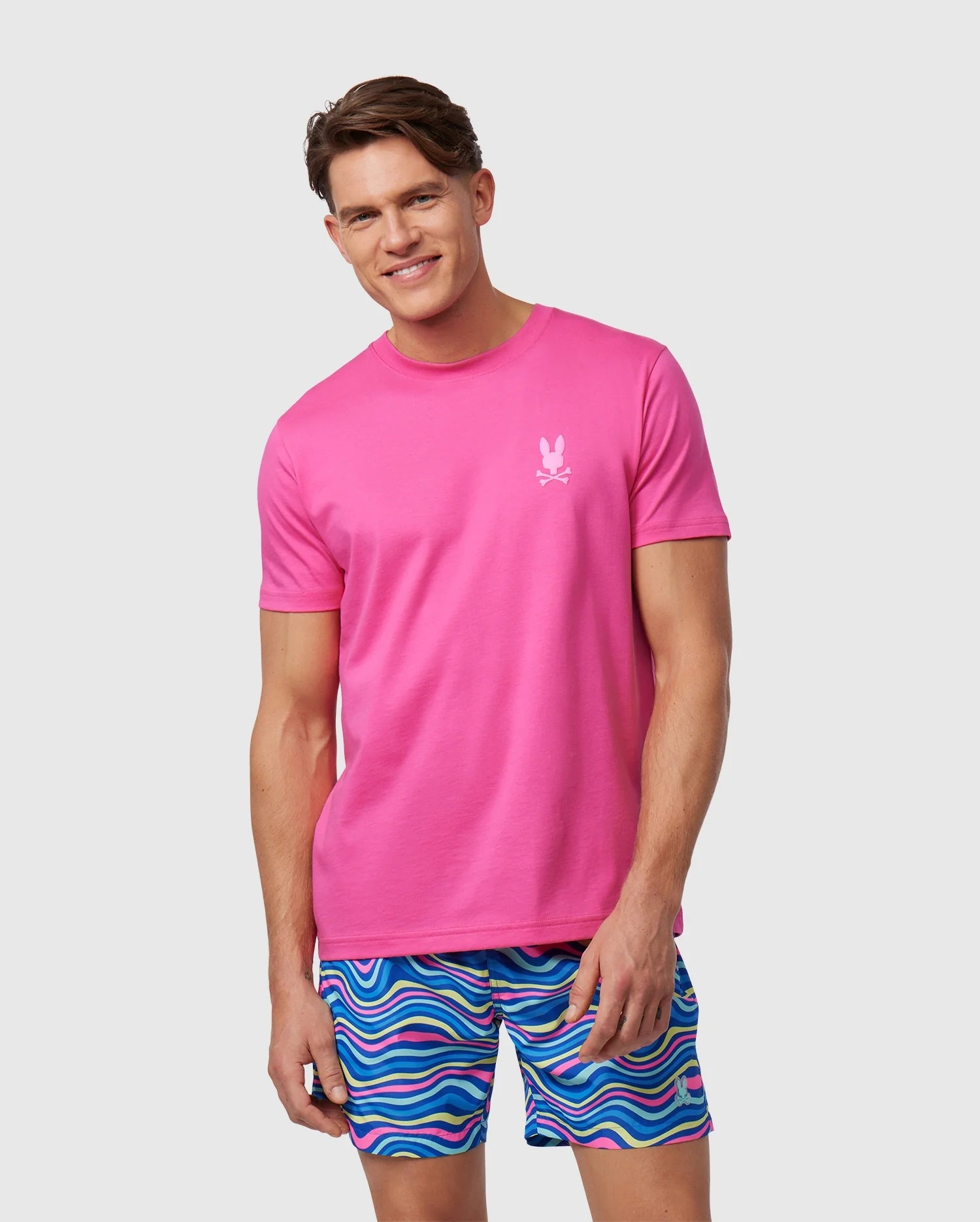 A man wearing a bright pink Psycho Bunny MENS MAYBROOK BACK GRAPHIC TEE (B6U196B2TS) with a small white logo on the chest and a back graphic, paired with colorful striped shorts, posing against a plain background.