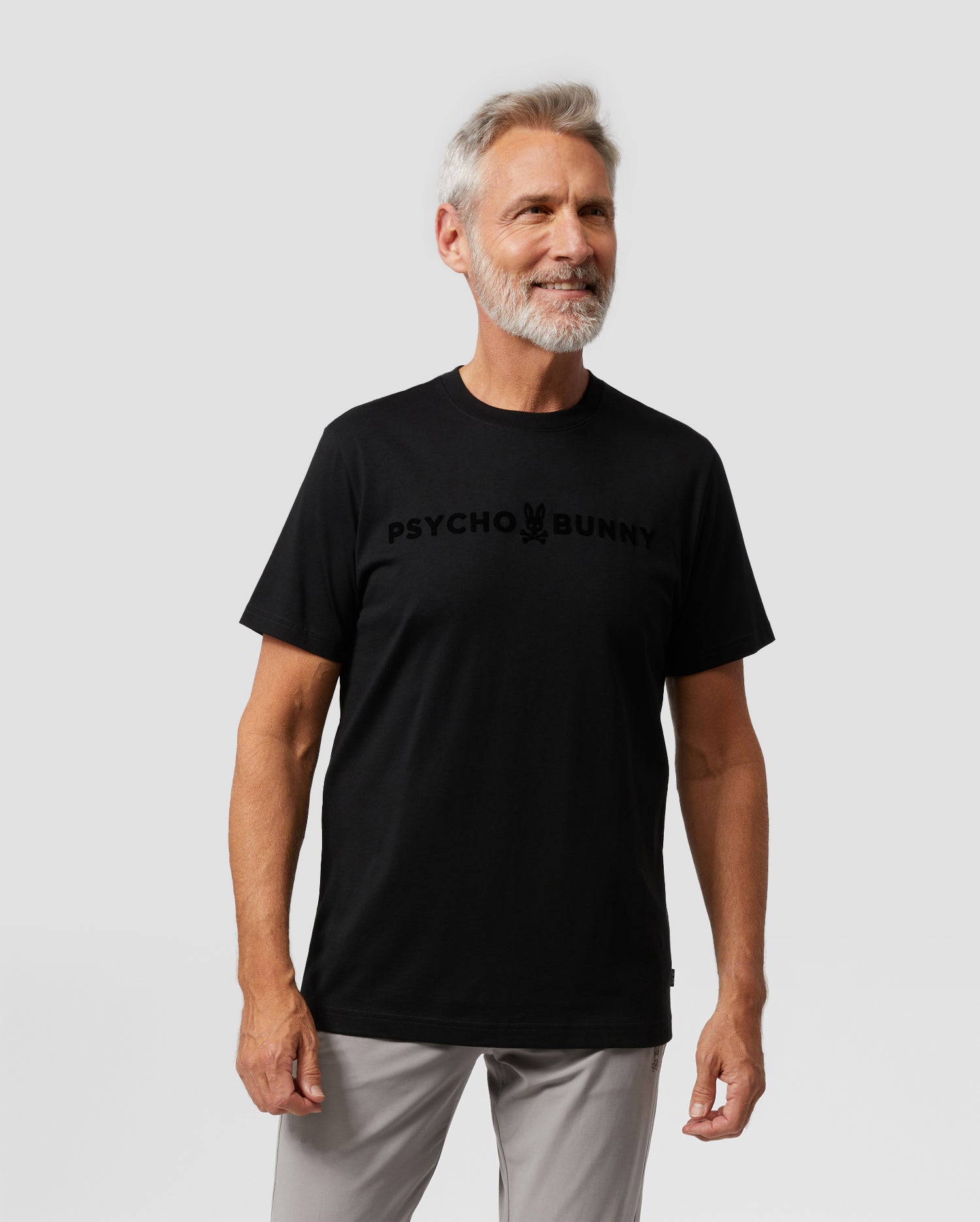 A man with short gray hair and a beard is posing against a plain gray background. He is wearing the Psycho Bunny MENS ROYCE FLOCKING GRAPHIC TEE - B6U248W1PC, crafted from Pima cotton jersey, with 