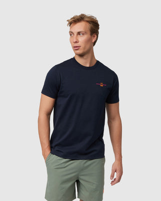 A man with short, light brown hair stands wearing a navy blue MENS WASTERLO BACK GRAPHIC TEE - B6U317B2TS by Psycho Bunny and light green shorts. He is looking off to his left with his left hand in his pocket. The background is plain white.