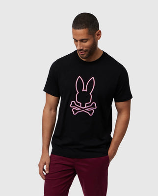 A man with short hair and a beard is standing and looking down. He is wearing a black Psycho Bunny MENS FLOYD GRAPHIC TEE - B6U338B2TS, made from Pima cotton, featuring an embroidered bunny outline and crossed bones design on the front. His outfit is completed with maroon pants against a plain gray background.