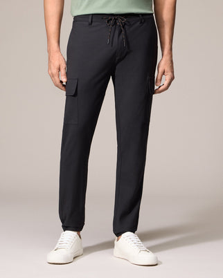 A person is wearing Psycho Bunny's MENS LLOYD CARGO JOGGER - B6P159B200 in black, complete with elasticized cuffs, standing against a neutral background. The pants feature utilitarian pockets and are paired with white sneakers and a light green top, with the photo focusing on the lower half of the body.